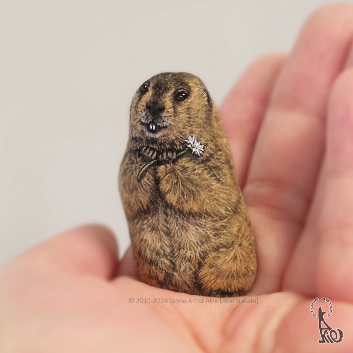 Hand-painted stone by Japanese artist resembling an adorable animal holding a small white flower in human palm.
