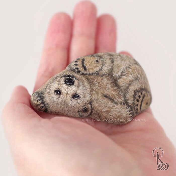Hand holding a stone painted by a Japanese artist to look like an adorable curled-up bear with detailed fur patterns.
