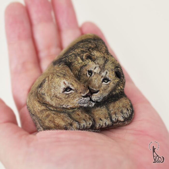 Hand holding a stone painted by Japanese artist, transformed into adorable animals resembling two cuddling lions.