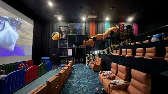 Indoor kids play area with slides and seating next to a movie screen showing an animated character, genius solutions setup.