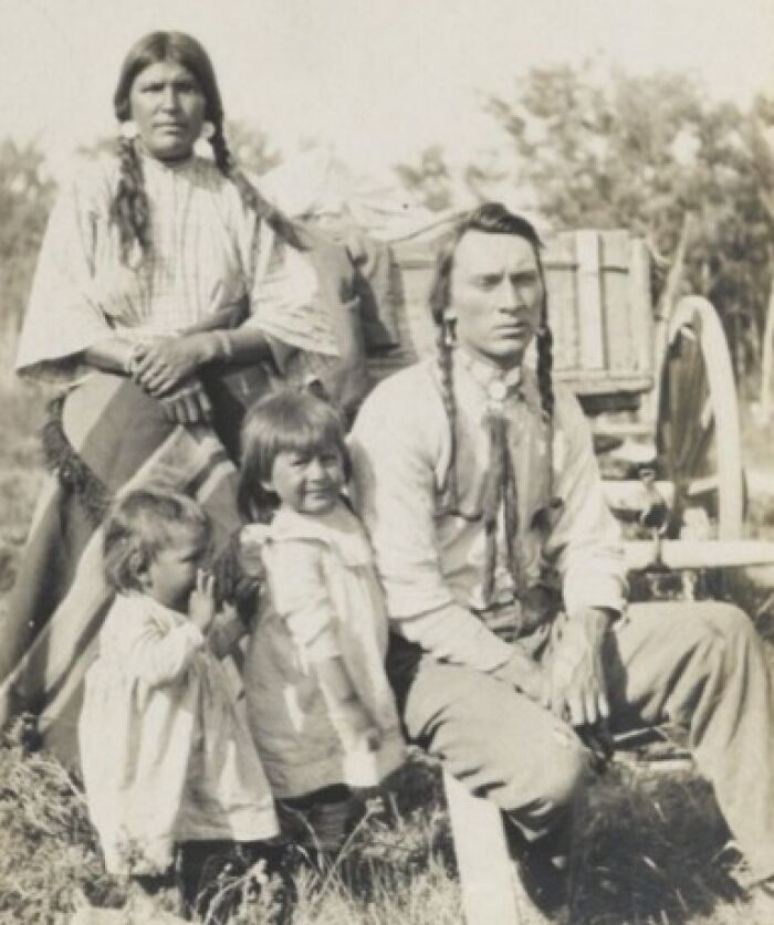Vintage Native American family portrait outdoors with parents and children dressed in traditional clothing near a wagon.