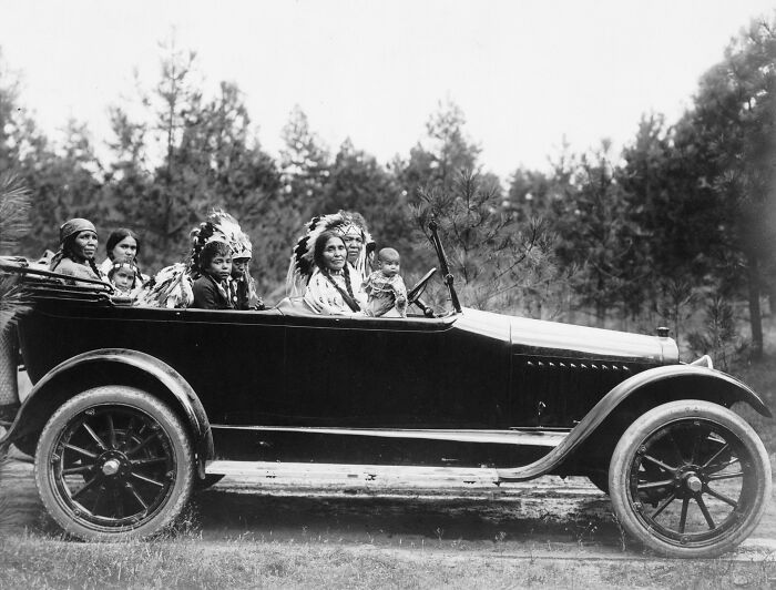 Vintage Native American family dressed in traditional attire riding in an early 20th-century open car outdoors.