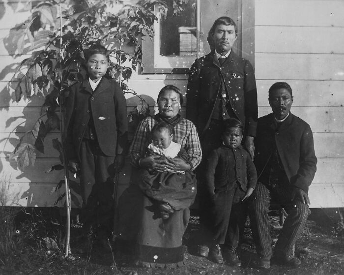 Vintage Native American family photo showing multiple generations posed outside a house in early 1900s traditional clothing.