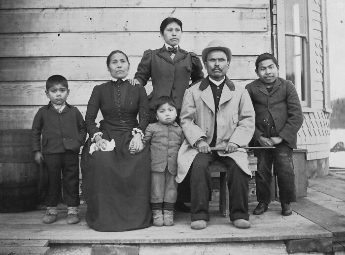 Vintage Native American family portrait showing adults and children dressed in early 20th-century clothing on a wooden porch.
