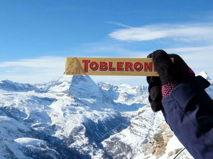 Hand wearing glove holding Toblerone chocolate bar with snowy mountain landscape in world geography setting.