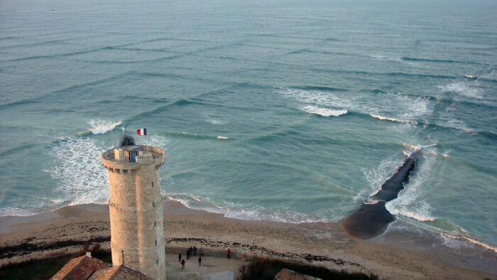 A coastal lighthouse overlooking the ocean with visible underwater geography and waves along the shore.