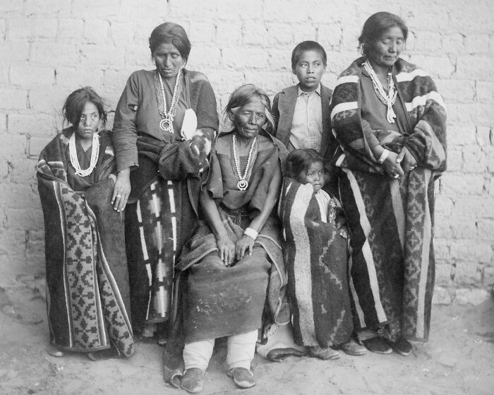Vintage Native American family portrait with traditional clothing and blankets, showing multiple generations together outdoors.