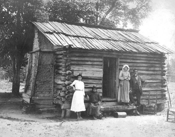Vintage Native American family posing outside a rustic log cabin surrounded by trees in a historic black and white photo.