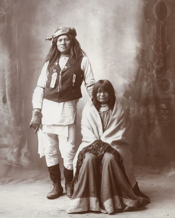 Vintage Native American family portrait with man standing and woman seated, both dressed in traditional early 20th century clothing.