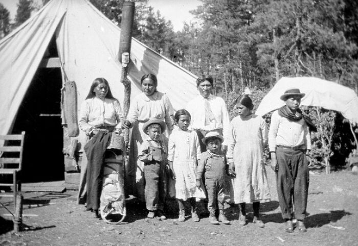 Vintage Native American family standing outside a tent in traditional and early 20th-century clothing in a forested area.