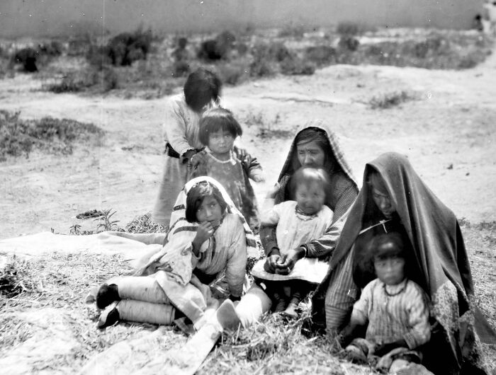 Vintage Native American family sitting outdoors in traditional clothing, capturing a moment of daily life in black and white.