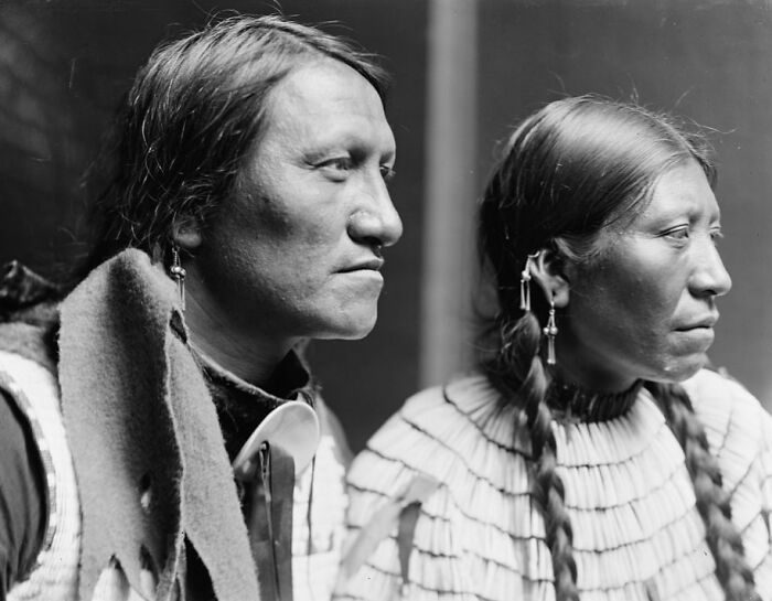 Vintage Native American family portrait showing a man and woman in traditional clothing and accessories, side profile view.