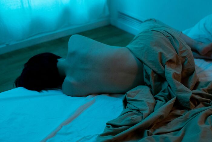 Person lying on bed covered with blanket in a dimly lit room, evoking peaceful neighbor tales and quiet woods living.