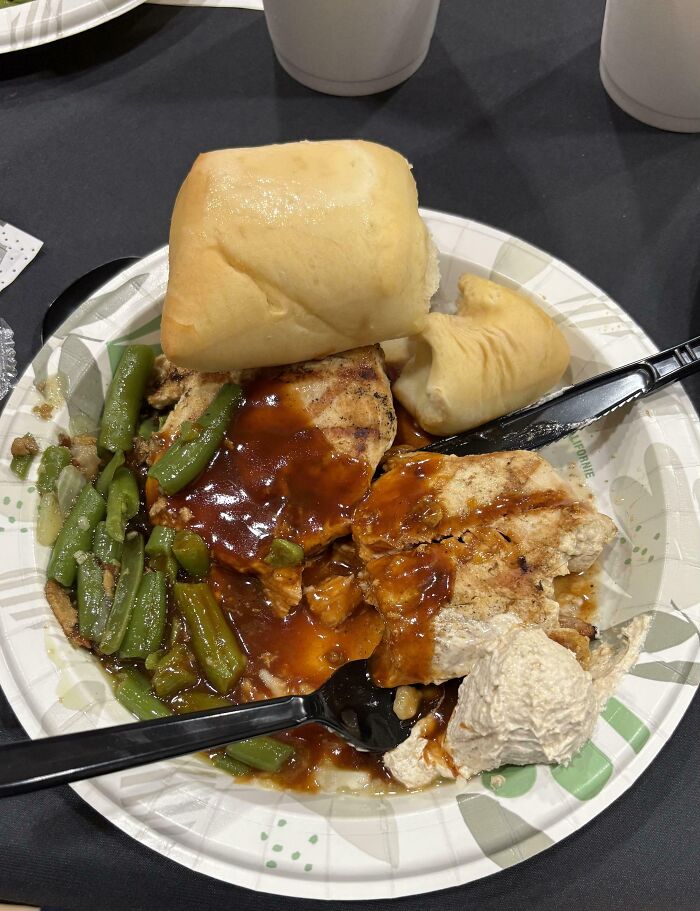 School cafeteria meal with green beans, grilled chicken covered in brown sauce, bread rolls, and a salad scoop on a disposable plate.
