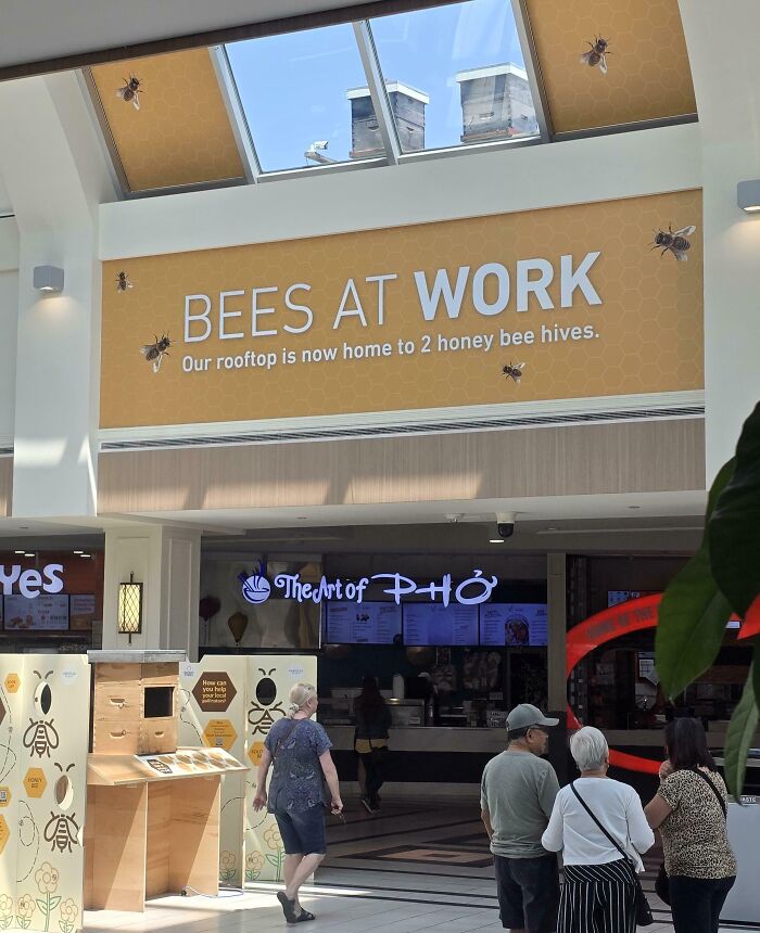 Indoor mall area with people walking near a bees at work sign promoting rooftop honey bee hives as a genius solution.