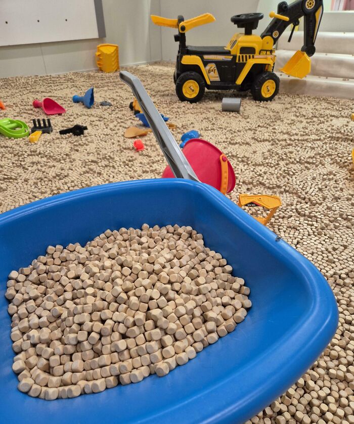 Blue wheelbarrow filled with small wooden pellets surrounded by toy construction vehicles and scattered tools on floor.