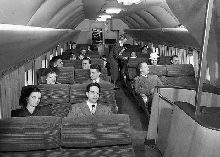 Passengers seated inside a 1950s airplane cabin with a flight attendant serving drinks, capturing life in the 1950s.