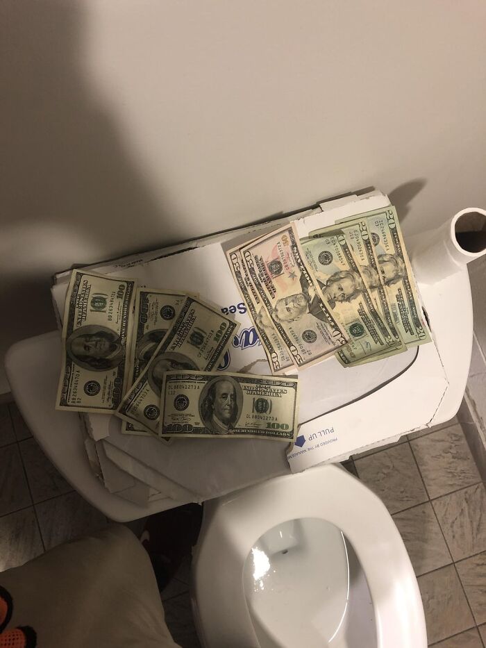 Money in various denominations placed on a closed pizza box on a toilet seat in a bathroom setting.