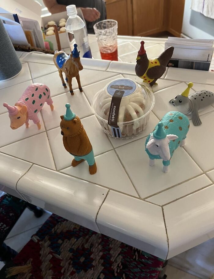 Assorted animal figurines wearing party hats gathered around a frosted cinnamon roll on a tiled kitchen counter.