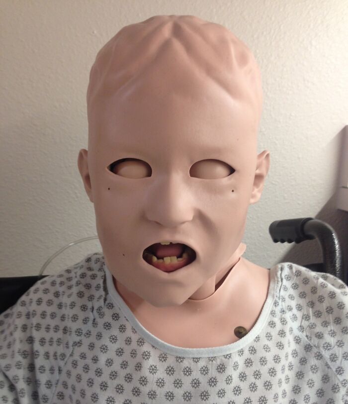 Medical mannequin with distorted facial features and blank eyes wearing a hospital gown in a clinical setting.