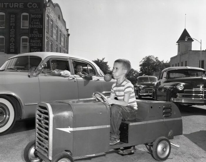 Young boy riding a pedal car and blowing a bubble in a classic 1950s Florida street scene with vintage cars around.