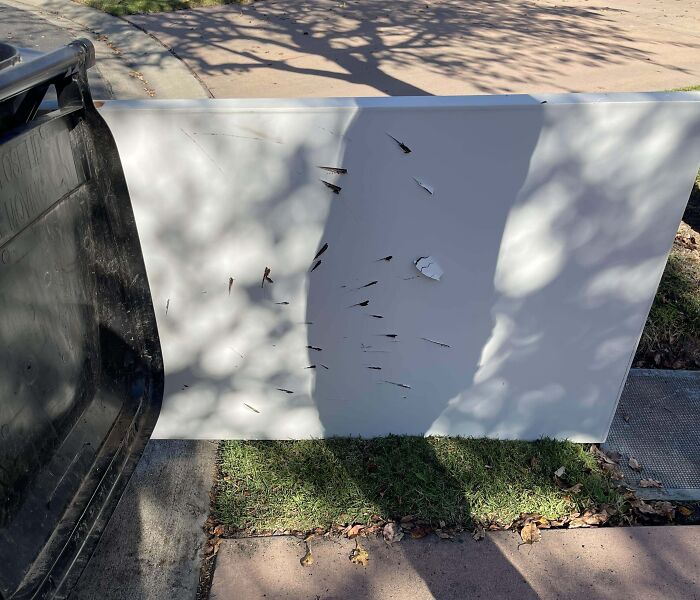 White panel with multiple deep puncture marks leaning against a trash bin on a sidewalk in a suburban neighborhood.