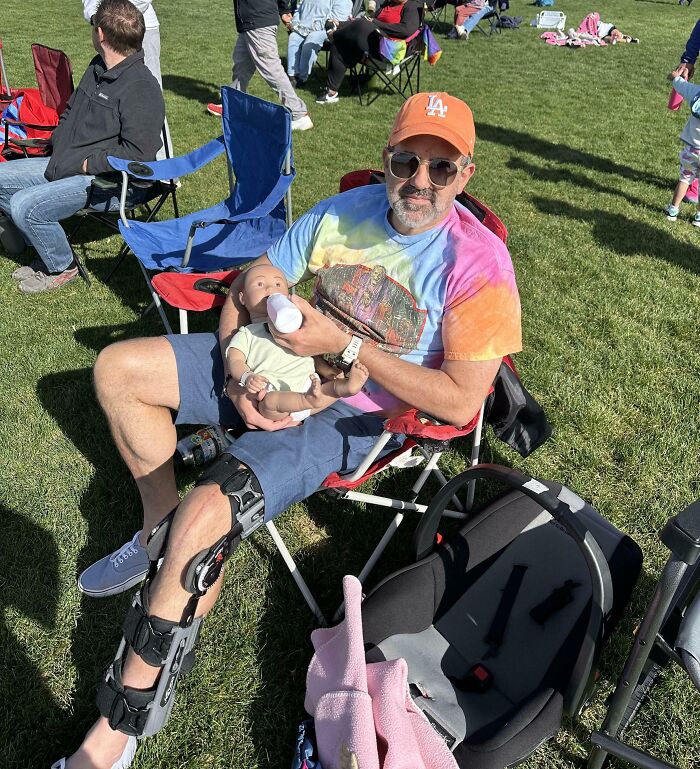 Dad wearing a leg brace and colorful shirt, sitting outdoors feeding baby with a bottle, showcasing hilarious dads nailing fatherhood.