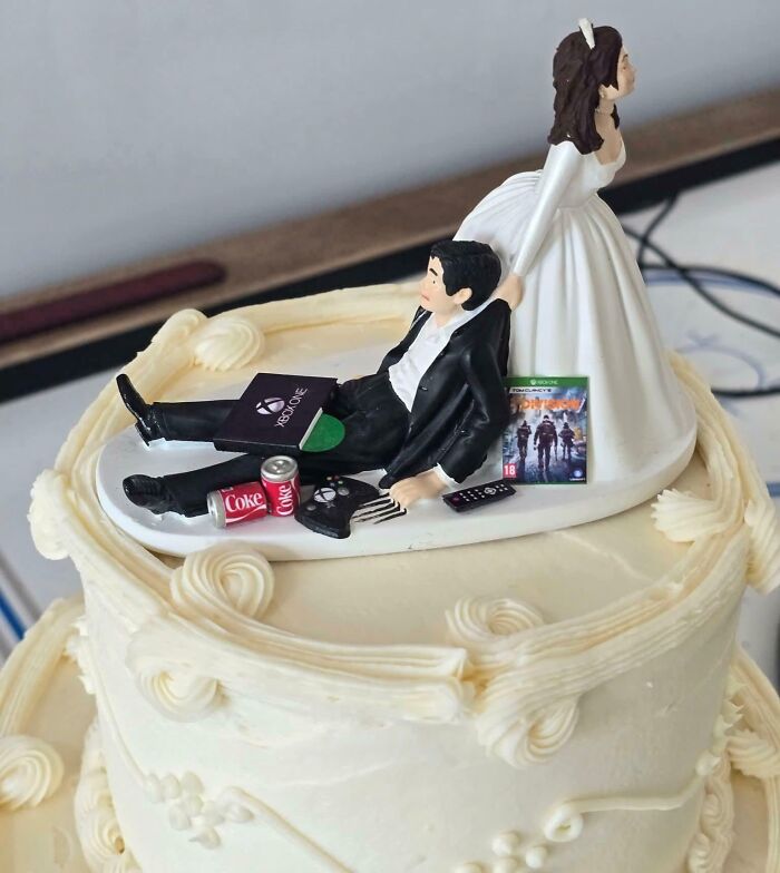 Bride figurine dragging groom figurine lying on wedding cake with video games, soda cans, and remote, showing groom entitlement.