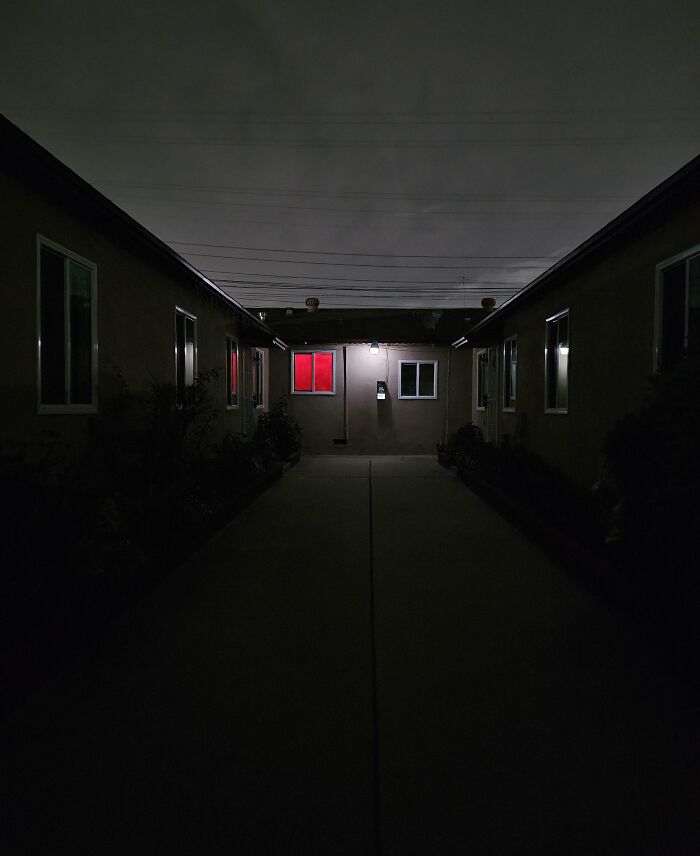 Dimly lit residential walkway at night with one window glowing red, evoking neighbor tales that make you want to move to the woods.