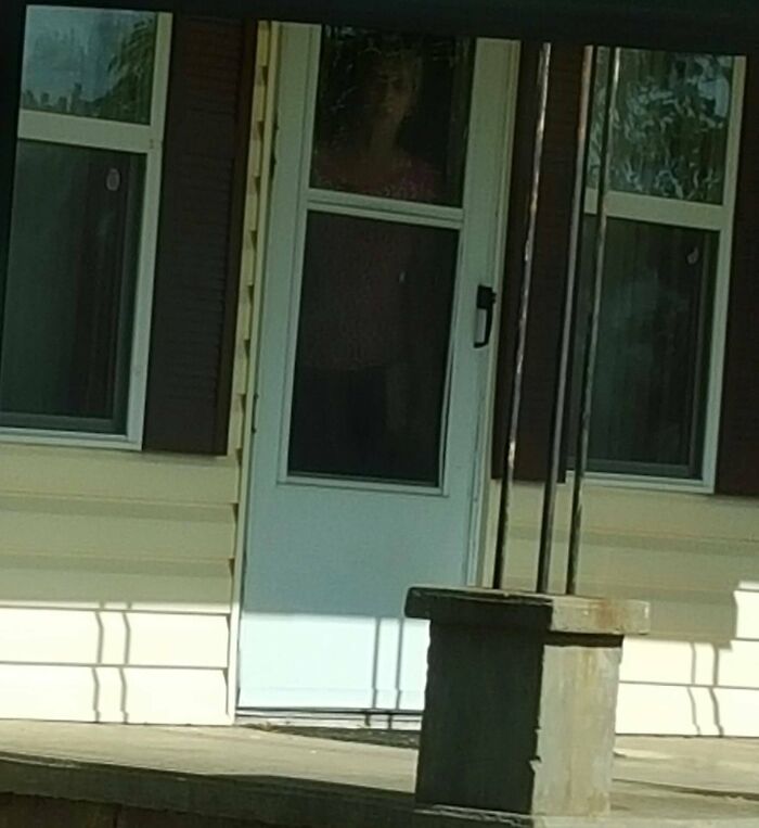 Person standing behind a screen door on a porch of a house, hinting at neighbor tales that inspire a move to the woods