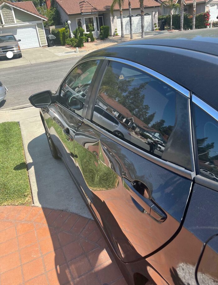 Black car with a large dent parked in a suburban neighborhood reflecting nearby houses and greenery.