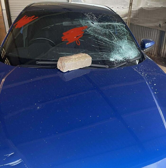 Blue car with a c*****d windshield and a brick on the hood, illustrating unusual neighbor tales from the woods.