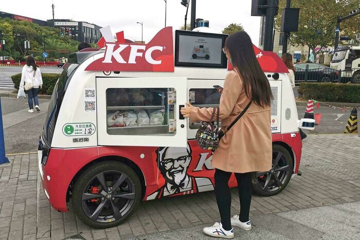 Autonomous KFC vending vehicle serving food to a woman on a city sidewalk, showcasing small genius country innovations.