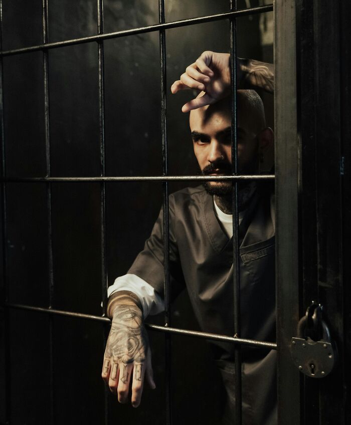 Man behind bars with tattoos on hand, looking inside dim cell, evoking intense neighbor tales and desire to move to the woods.