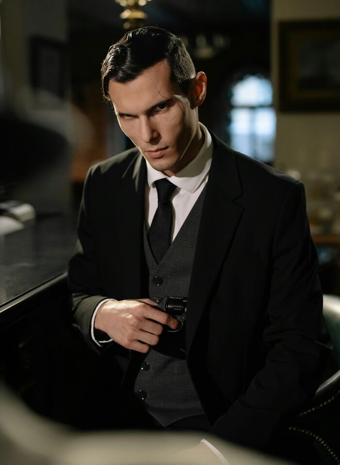 Young man in a dark suit holding a gun indoors, evoking suspenseful neighbor tales that might make you want to move to the woods.