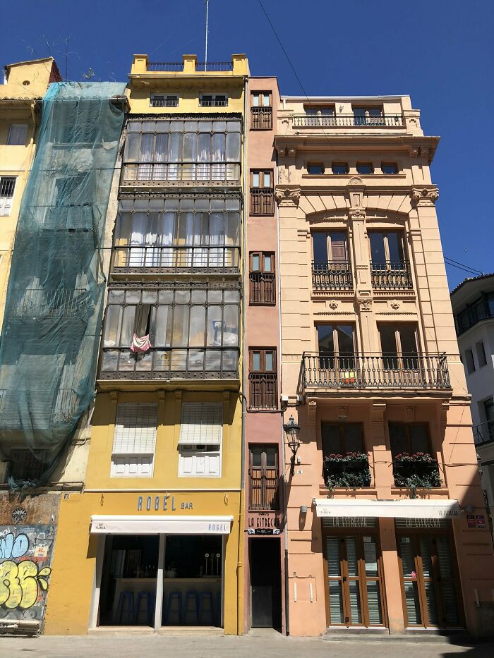 Narrow building squeezed between two wider buildings, showcasing unique small architectural design in urban setting under clear blue sky.