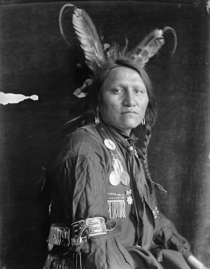 19th century photo of a Native American man wearing traditional clothing and feathered headdress, history captured in black and white.