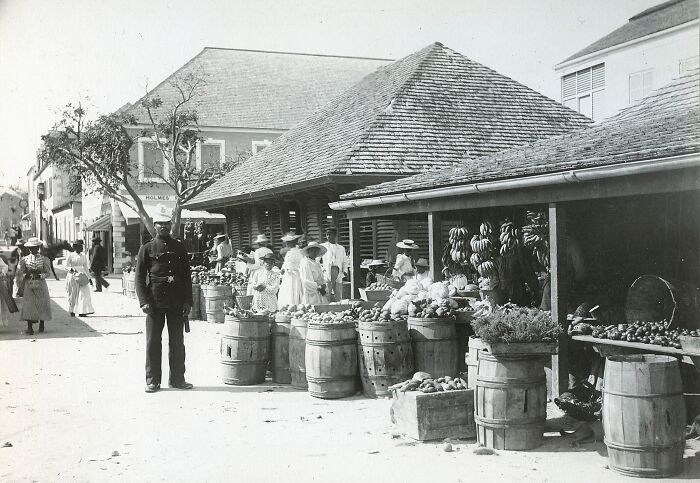 Black and white 19th century photo capturing a busy market street with people shopping and wooden barrels of produce.