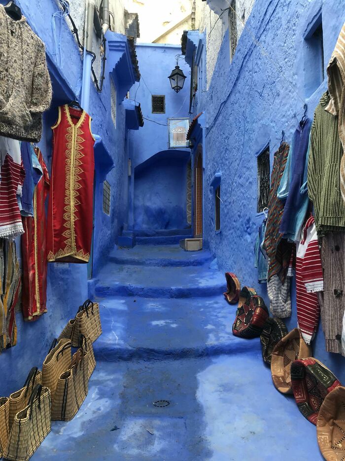 Narrow blue-painted alleyway with traditional clothing and woven bags displayed for sale in a vibrant market setting.