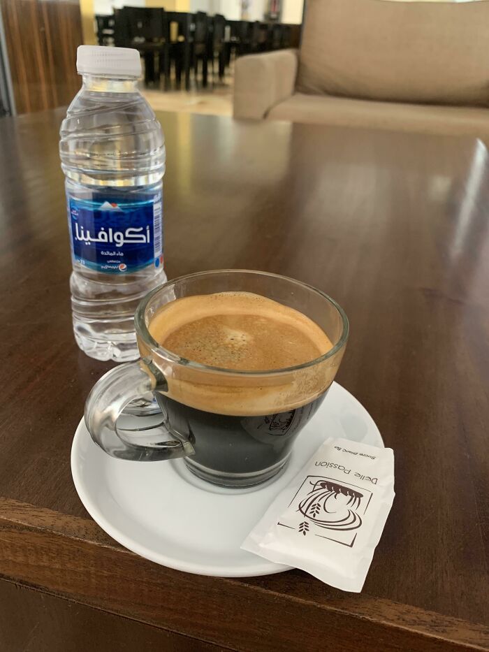 A cup of black coffee on a white saucer with sugar packet and a bottle of water, showcasing small genius country details.