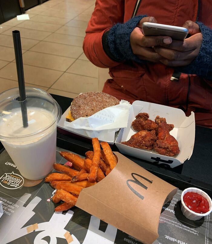 Person using smartphone with fast food tray featuring sweet potato fries, burger, wings, and milkshake, showcasing small genius food ideas.
