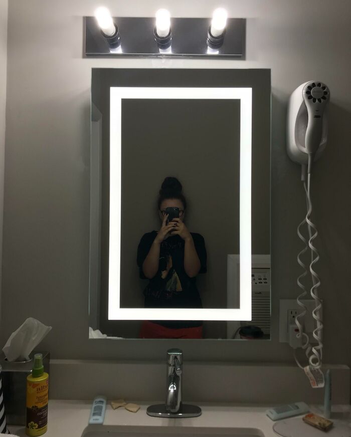 Hotel bathroom mirror with damaged lower corner lighting and a person taking a selfie in reflection, showing hotel mess up.