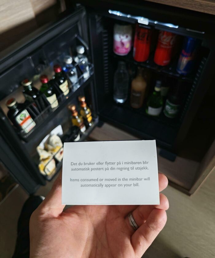 Hand holding a note about minibar charges in front of an open hotel minibar filled with drinks and snacks.