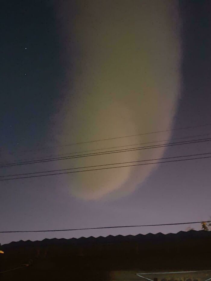 Unusual sky phenomenon glowing with a large pale cloud formation above rooftops and power lines at night.