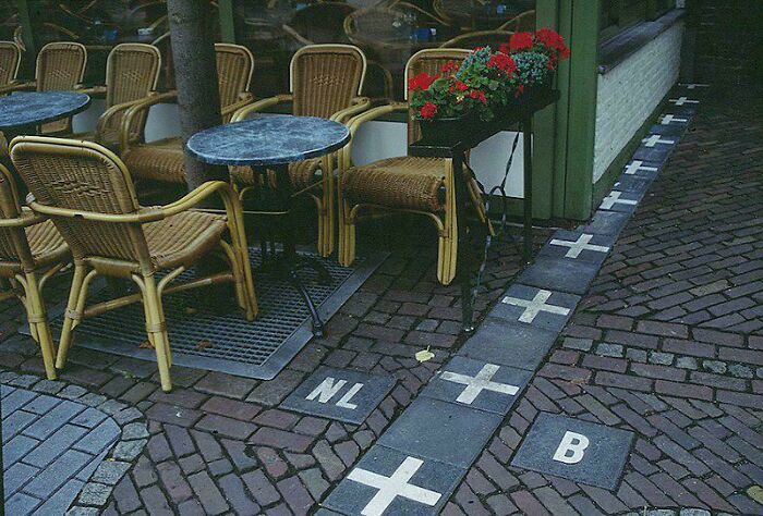 Outdoor café seating with chairs and tables beside a unique border line marked with crosses and country codes NL and B.