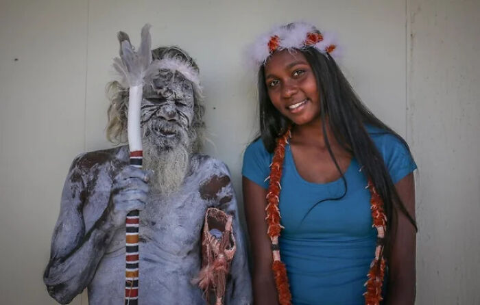 Elderly man with body paint and traditional staff standing next to young woman with floral headband and garland, extraordinary sight.