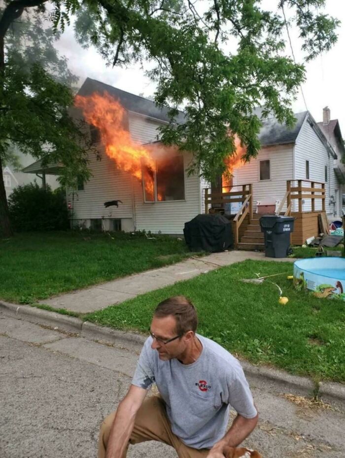 Man crouching near a house fire with flames coming from windows, an extraordinary and fascinating sight captured outdoors.