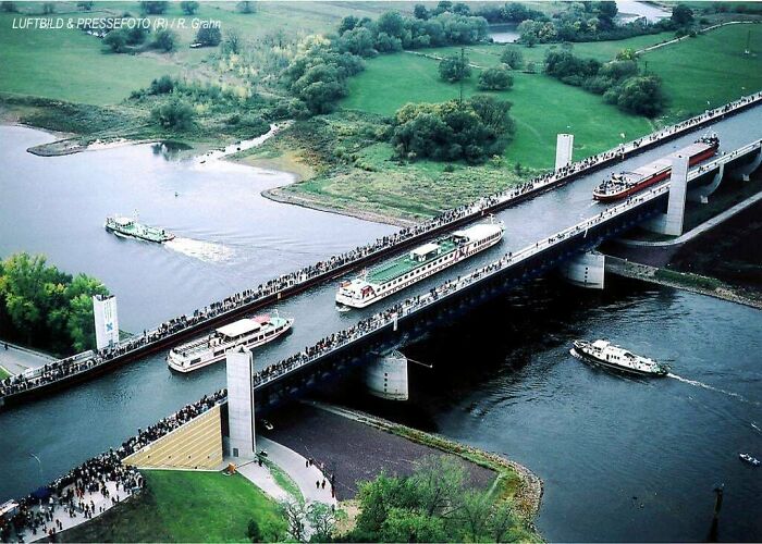 A fascinating sight of boats passing on an elevated water bridge crowded with people along the banks and green landscape.
