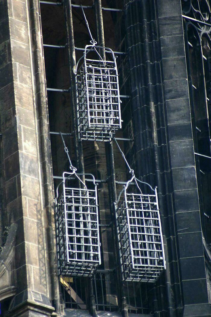 Three metal cages hanging on ropes against a dark stone building, an extraordinary and fascinating sight.