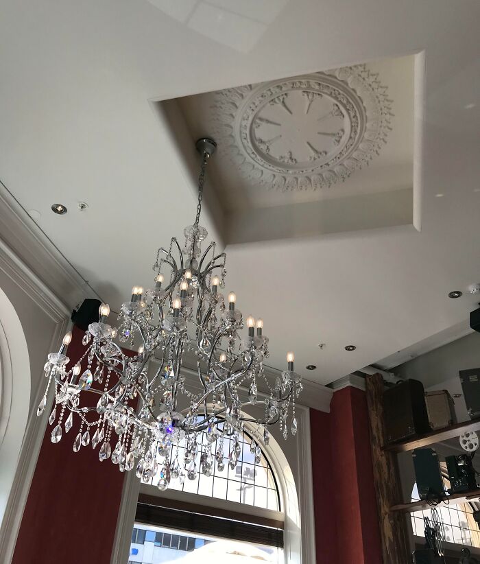 Crystal chandelier hanging below an ornate ceiling medallion in a room with large windows and vintage decor, highlighting imperfect details.